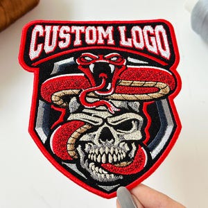 May include: A shield-shaped embroidered patch featuring a skull entwined with a red and tan snake. The top section displays the white text CUSTOM LOGO on a black background. The design is bordered by a thick red edge with black and grey accents.