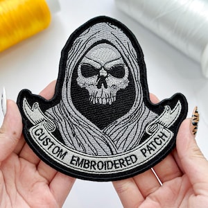 May include: Embroidered patch featuring a skull in a hooded cloak design. The patch is black with gray and white embroidery, and the text "CUSTOM EMBROIDERED PATCH" is displayed on a banner. This patch is suitable for jackets, bags, or other fabric items.