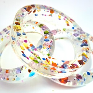 May include: Three clear resin bangles with colorful confetti-like inclusions. The bangles are linked together, showcasing a variety of colors including blue, orange, yellow, and purple. The resin is transparent, allowing the colors to be seen.