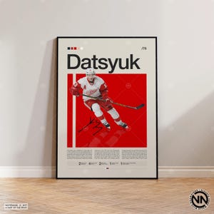 Könnte beinhalten: Ein gerahmtes Poster mit einem Eishockeyspieler in roter und weißer Uniform, mit dem Namen "Datsyuk" in großen schwarzen Buchstaben. Das Poster hat einen roten Hintergrund mit dem Bild und der Unterschrift des Spielers. Das Poster wird auf einem Holzboden angezeigt.