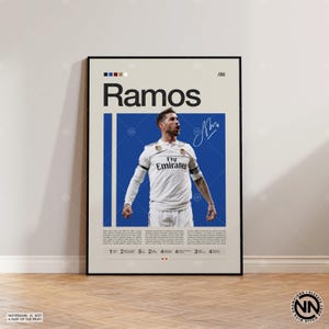 Op de afbeelding: Een ingelijste poster met de naam "Ramos" in zwart, boven een foto van een voetballer in een wit shirt. De poster bevat een blauwe verticale streep, een handtekening en de tekst "Fly Emirates."