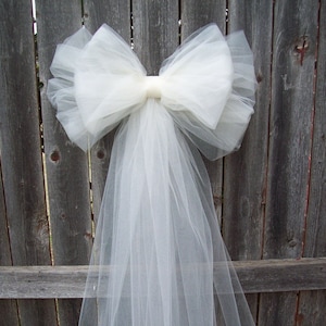 May include: A large, white tulle bow with long, flowing streamers. The bow is attached to a wooden fence. The tulle is a soft, sheer fabric, creating a delicate and airy appearance. This decorative bow is suitable for weddings or other special events.