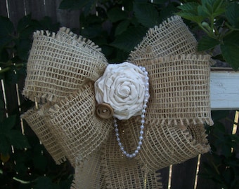May include: A large burlap bow with a white fabric flower and a pearl necklace. The bow is tied in a knot and the flower is centered in the middle of the bow.
