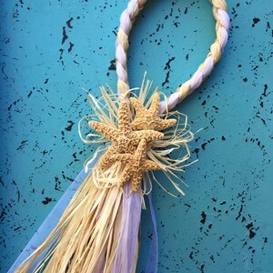 May include: A light purple and natural straw rope wreath with three starfish attached to the bottom. The wreath is hanging on a blue textured wall.