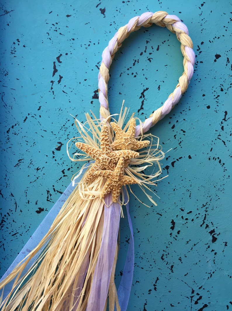 May include: A light purple and natural straw rope wreath with three starfish attached to the bottom. The wreath is hanging on a blue textured wall.