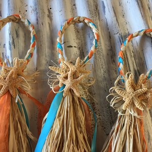 May include: Three woven rings with orange and turquoise ribbon accents, decorated with starfish and raffia. The rings are tied together with a bow.