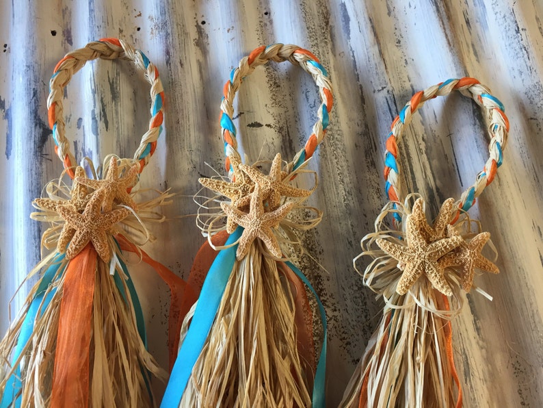 May include: Three woven rings with orange and turquoise ribbon accents, decorated with starfish and raffia. The rings are tied together with a bow.