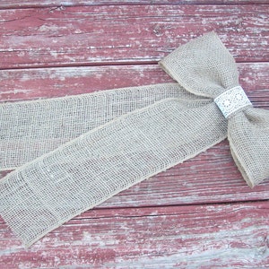 May include: A large burlap bow with a white lace accent. The bow is tied with a long tail and is lying on a weathered red wooden surface.