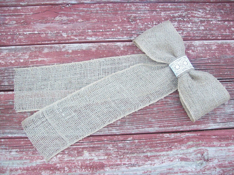 May include: A large burlap bow with a white lace accent. The bow is tied with a long tail and is lying on a weathered red wooden surface.