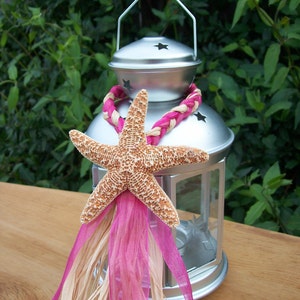 May include: A silver lantern with a starfish and pink and beige ribbon decoration. The lantern has a glass window and a handle on top.