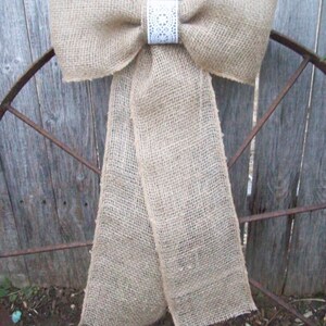 May include: A large burlap bow with a white lace center, tied to a rusty metal fence.