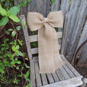 May include: A large, rustic burlap bow tied on a wooden chair. The bow is a light brown color and has a natural, textured look.