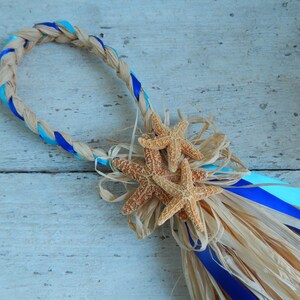 May include: A braided wreath made of natural raffia with blue and turquoise ribbon accents. The wreath is decorated with three starfish and a raffia tassel.