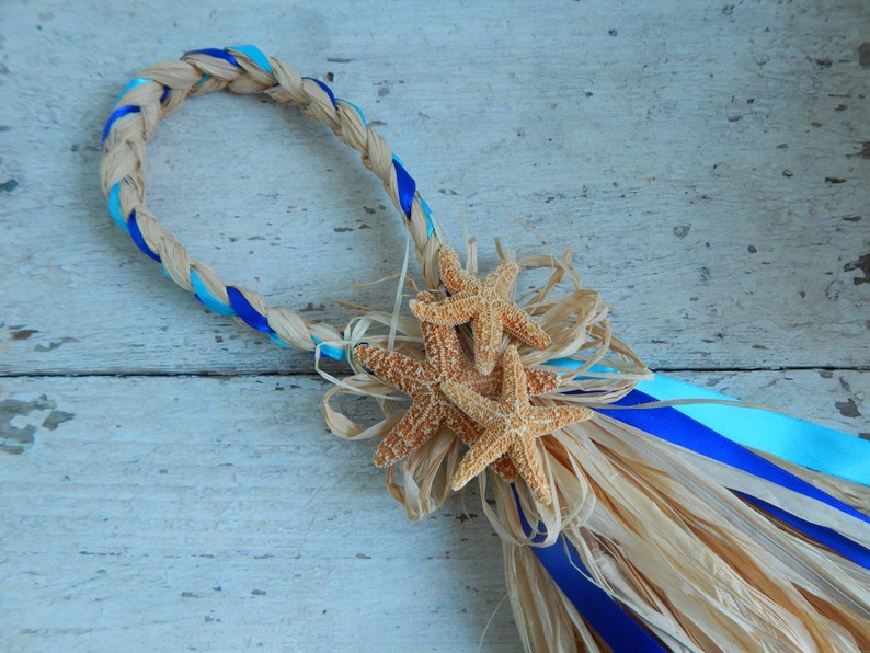 May include: A braided wreath made of natural raffia with blue and turquoise ribbon accents. The wreath is decorated with three starfish and a raffia tassel.