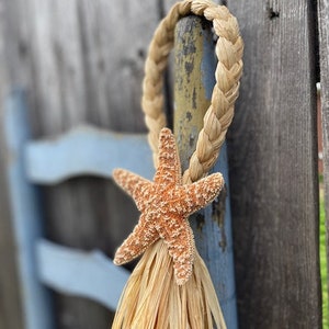 May include: A natural fiber wreath with a starfish attached. The wreath is made of woven straw and has a long, fringed tassel hanging from the bottom.