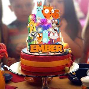 Op de afbeelding: Een verjaardagstaarttopper met meerdere niveaus, voorzien van een kleurrijke reeks stripfiguren, een discobal en de naam EMBER in dikke oranje letters. De topper is verlicht met kleine lampjes en staat bovenop een taart met brandende kaarsjes.
