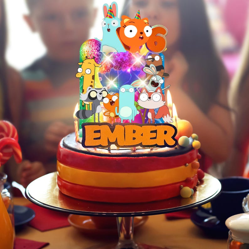 Op de afbeelding: Een verjaardagstaarttopper met meerdere niveaus, voorzien van een kleurrijke reeks stripfiguren, een discobal en de naam EMBER in dikke oranje letters. De topper is verlicht met kleine lampjes en staat bovenop een taart met brandende kaarsjes.