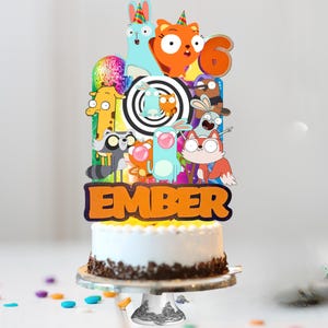 Op de afbeelding: Een taarttopper met een kleurrijke collage van stripfiguren, een discobal, een zwart-witte spiraal, het cijfer 6 en de naam Ember in oranje letters, geplaatst op een wit geglazuurde taart met chocoladevlokken.
