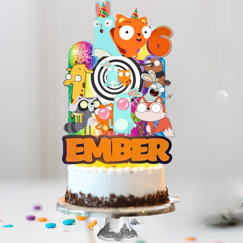 Op de afbeelding: Een taarttopper met een kleurrijke collage van stripfiguren, een discobal, een zwart-witte spiraal, het cijfer 6 en de naam Ember in oranje letters, geplaatst op een wit geglazuurde taart met chocoladevlokken.