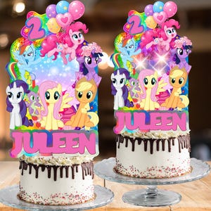 May include: Two round cakes with white frosting and dark chocolate ganache drips, topped with large, colorful My Little Pony character cutouts, balloons, the number 2, and the name JULEEN in bold pink letters on a holographic background.