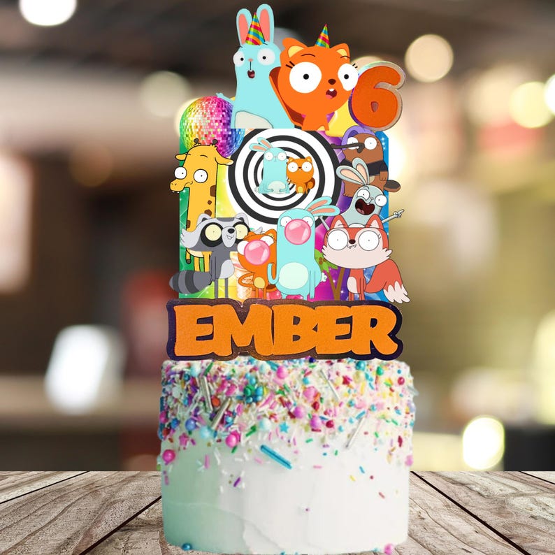 Op de afbeelding: Een gepersonaliseerde taarttopper met stripfiguren, een discobal en een zwart-wit spiraalpatroon. De topper toont de naam EMBER in grote oranje letters en het cijfer 6, geplaatst op een witte taart met kleurrijke hagelslag.