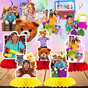 May include: A collection of party decorations featuring various cutouts of people, teddy bears, and cartoon characters, including a red boombox, all mounted on yellow and red paper honeycomb bases, arranged on a wooden surface against a purple background.