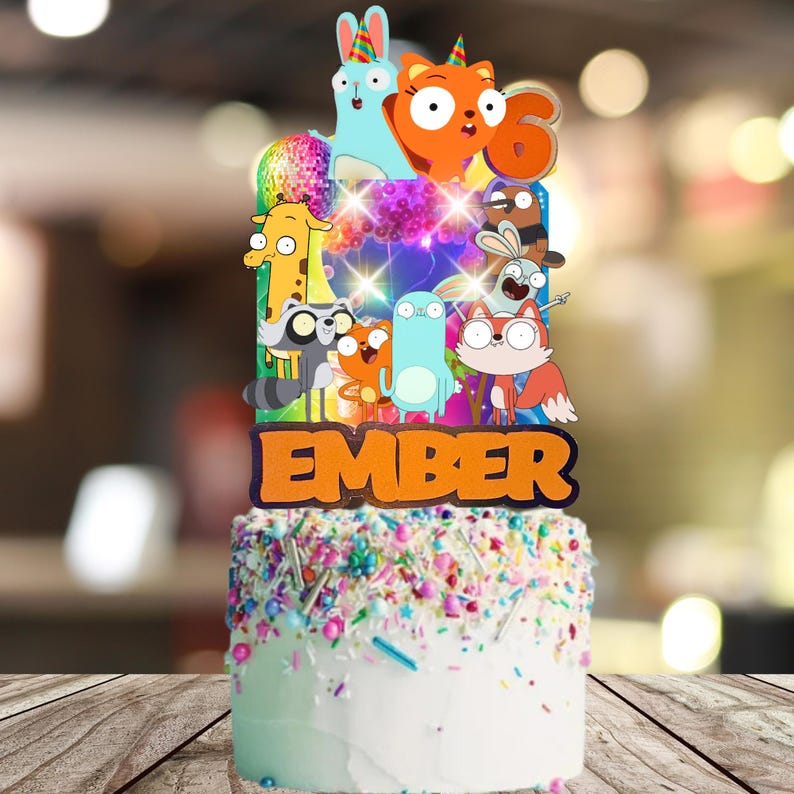 Op de afbeelding: Een taarttopper met een kleurrijke reeks cartoon-dierfiguren, een discobal en het cijfer zes, met de naam EMBER in grote oranje letters aan de basis. De topper is geplaatst in een witte taart die bedekt is met veelkleurige hagelslag en decoraties.