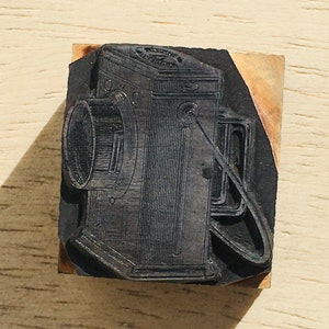 May include: A vintage wooden printing block featuring a detailed illustration of a camera. The camera is black and white and has a lens with a circular shape. The block is made of wood and has a rough texture.