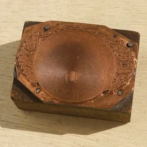 May include: A vintage copper printing block with a circular design and intricate details. The block is mounted on a wooden base and has a worn, aged appearance.
