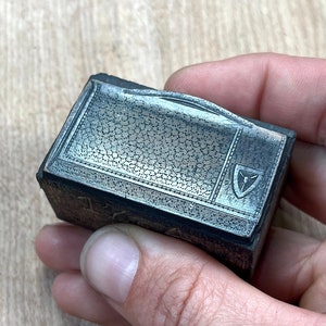 May include: A small, silver-coloured metal box with a textured lid and a small, raised shield-shaped design on the front. The box is approximately 7.5 cm long and 5 cm wide.