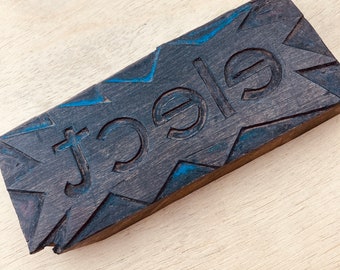 Puede incluir: Un bloque de madera oscura con la palabra "Jole" tallada en él. Las letras están delineadas con pintura azul y el fondo es un patrón de veta de madera.