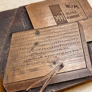 May include: A vintage metal printing block featuring the text "W3M Solid Rubber Type" and a range of letters and numbers. The block is made of brass and has a worn, aged look.
