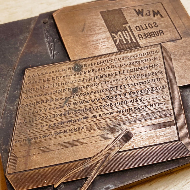 May include: A vintage metal printing block featuring the text "W3M Solid Rubber Type" and a range of letters and numbers. The block is made of brass and has a worn, aged look.