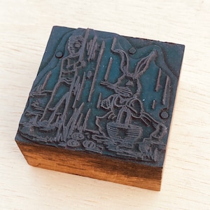 Pu&ograve; includere: Un timbro da stampa in legno vintage con un'illustrazione in bianco e nero di un coniglio e una persona che tiene un bastone. Il timbro &egrave; in legno e ha una finitura marrone scuro.