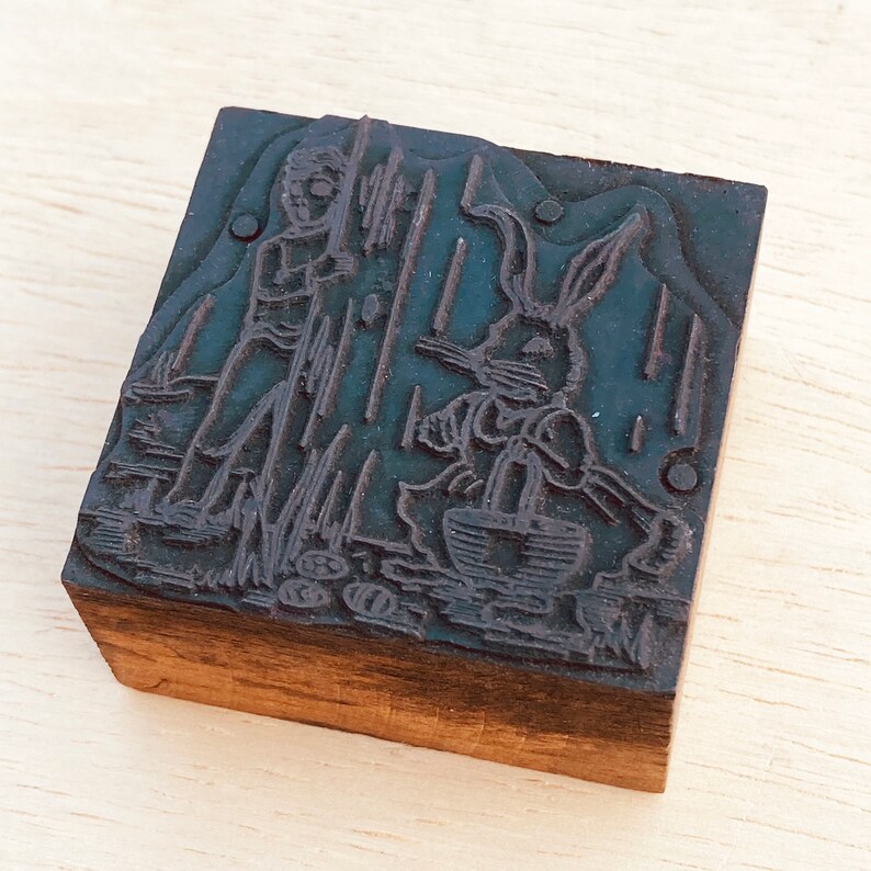 Pu&ograve; includere: Un timbro da stampa in legno vintage con un'illustrazione in bianco e nero di un coniglio e una persona che tiene un bastone. Il timbro &egrave; in legno e ha una finitura marrone scuro.