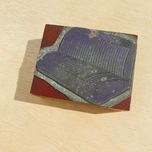 May include: A close-up of a vintage printing block with a raised image of a car seat. The seat is made of a textured fabric with a diamond pattern and is a dark purple colour.