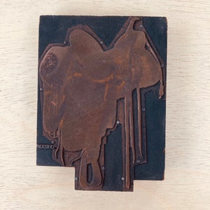May include: A vintage wooden printing block with a copper-coloured design of a saddle. The block has a black background and is slightly worn.