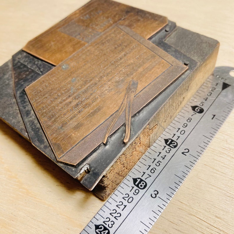 May include: A close-up of a metal printing block with a raised design. The block is mounted on a wooden base and is being measured with a ruler. The ruler shows measurements in inches.