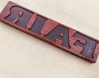 Puede incluir: Un letrero de madera con la palabra "FAIR" tallada. Las letras están pintadas de negro y la madera es de color marrón rojizo oscuro.