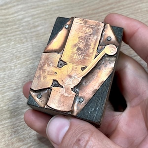 May include: A vintage wood and copper printing block with a design of a kerosene lamp. The lamp is labeled "Oil" and has a wick and a handle. The block is held in a hand.