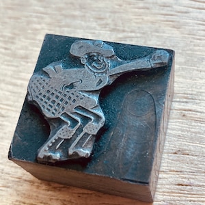 May include: A vintage metal printing block featuring a cartoon figure wearing a hat and riding a horse. The block is made of dark gray metal and has a worn, textured surface.