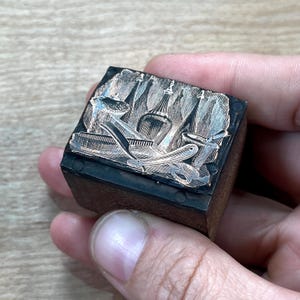 May include: A vintage wood printing block with a design of a barber's tools, including razors, combs, and a shaving brush. The design is engraved in the wood and has a silver finish.