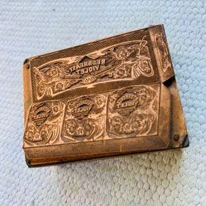 Puede incluir: Un bloque de impresi&oacute;n de madera vintage con las palabras "VIOLET REDBREAST" y "VIOLET" en relieve. El bloque tiene un dise&ntilde;o floral decorativo y est&aacute; hecho de madera marr&oacute;n claro.