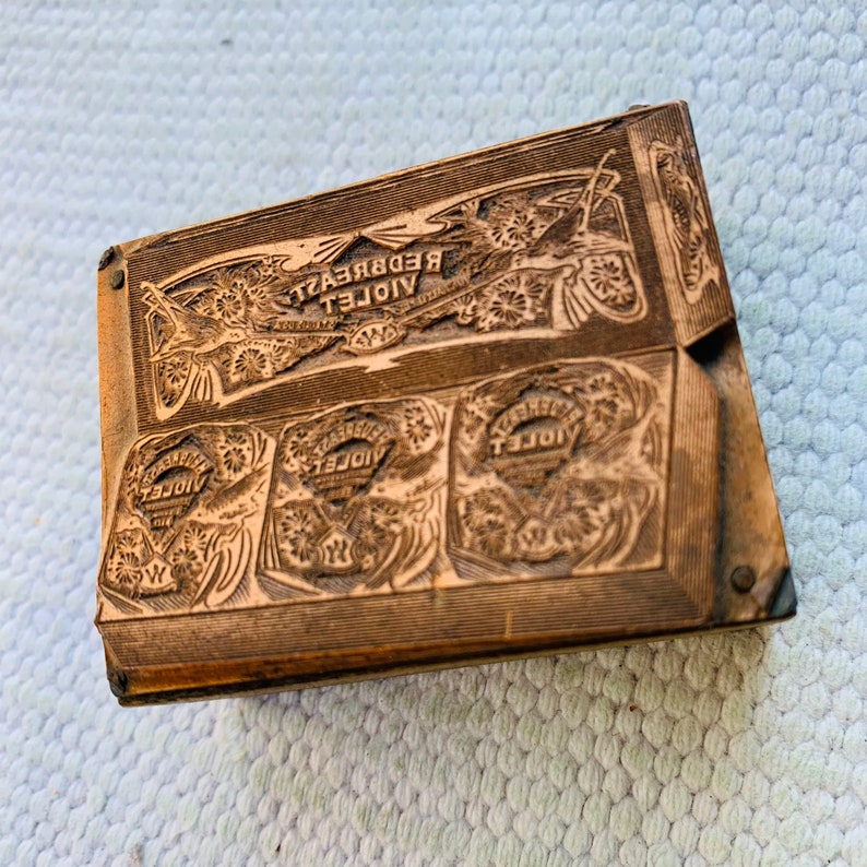 Puede incluir: Un bloque de impresi&oacute;n de madera vintage con las palabras "VIOLET REDBREAST" y "VIOLET" en relieve. El bloque tiene un dise&ntilde;o floral decorativo y est&aacute; hecho de madera marr&oacute;n claro.