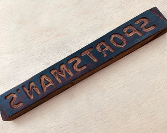 May include: A wooden block with the word "SPORTSMAN'S" carved into it in reverse lettering.