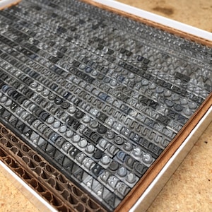 May include: A close-up of a metal printing block set in a white box. The blocks are arranged in rows and columns, and they feature a variety of letters and numbers. The blocks are made of a silver-coloured metal and have a slightly textured surface.