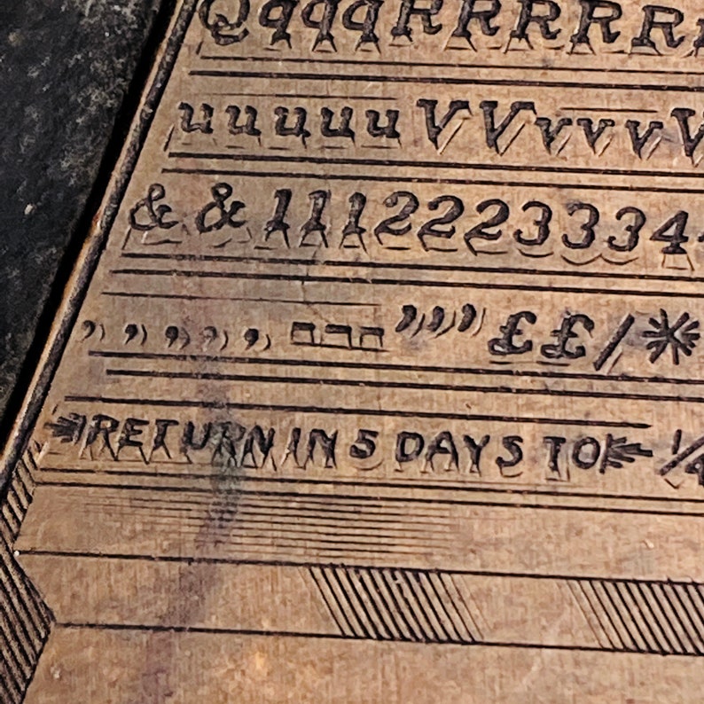 May include: Close-up of a wooden surface with engraved letters, numbers, and symbols. The text reads "RETURN IN 5 DAYS TO" followed by a symbol.