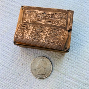 Puede incluir: Un bloque de impresi&oacute;n vintage de cobre con las palabras "VIOLET REDBREAST" y "VIOLET" en relieve. El bloque tiene un dise&ntilde;o floral decorativo y es aproximadamente del tama&ntilde;o de una moneda de euro.