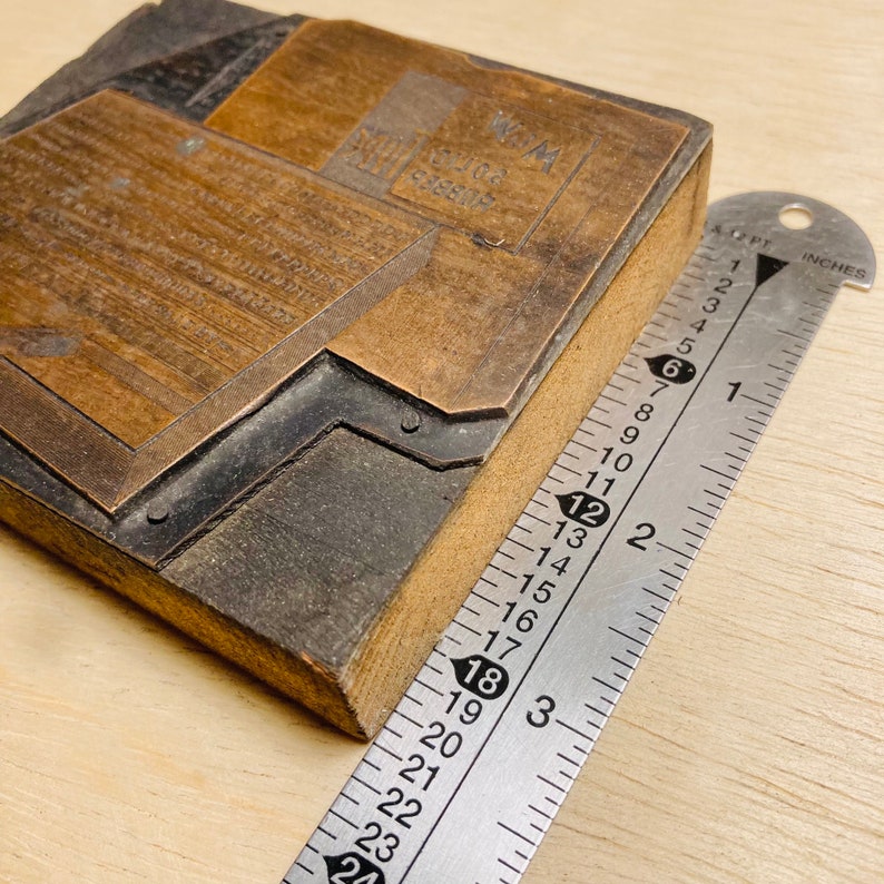 May include: A close-up of a wooden printing block with a metal plate. The plate has the text "WM. RICE & CO. CEREALS" engraved on it. The block is being measured with a ruler, which shows that it is approximately 7.6 cm wide.