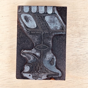 Puede incluir: Un bloque de impresión de metal negro y plateado con un diseño de una figura humana estilizada con un ojo grande y una serie de líneas y formas. El texto "1 2 3 4 Knife edge" es visible en el bloque.